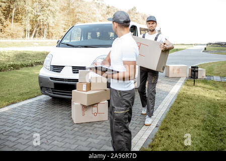 Two male couriers delivering goods by cargo van vehicle, unloading cardboard parcels from a car and checking list outdoors Stock Photo