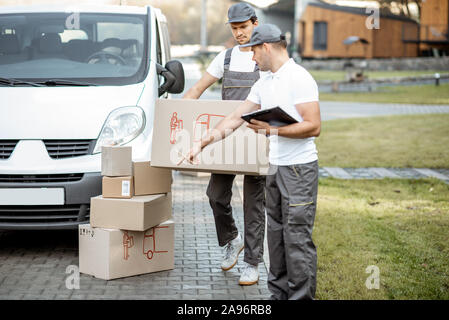 Two male couriers delivering goods by cargo van vehicle, unloading cardboard parcels from a car and checking list outdoors Stock Photo