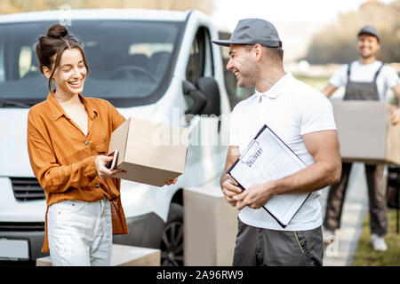 Courier with checklist delivering goods to a young woman by cargo van vehicle, mover with cardboard parcels on the background Stock Photo