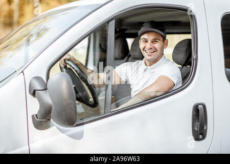 Portrait of a cheerful delivery driver in uniform looking out the window of the white cargo van vahicle, delivering goods by car Stock Photo