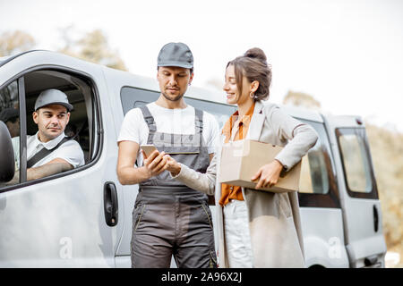 Delivery company employees in uniform delivering goods to a client by cargo van vehicle, woman signing on a smartphone, receiving parcel outdoors Stock Photo