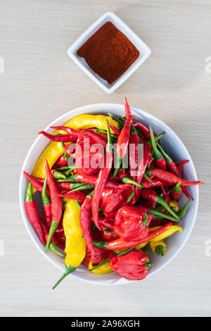 Mixed fresh hot chili peppers in porcelain bowl and ground peppers in small jar on a wooden table viewed from above Stock Photo