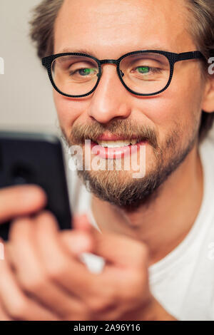 Handsome smiling young man with glasses lying on bed while holding his mobile phone. Stock Photo
