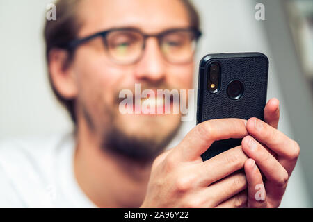 Handsome smiling young man with glasses lying on bed while holding his mobile phone. Stock Photo