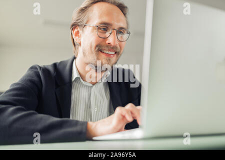 Photo of a handsome focused and smiling young man sitting by the desk working at the computer. Stock Photo