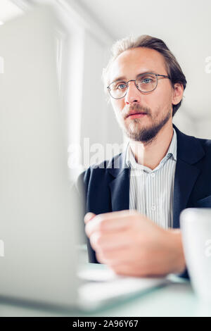 Photo of a handsome focused young man sitting by the desk and working at the computer. Stock Photo