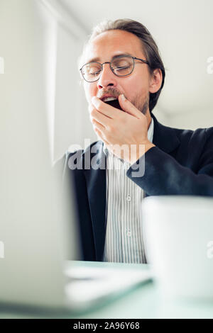 Tired young handsome man is yawning while working at the computer. Coffee cup visible next to him. Stock Photo