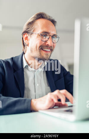 Photo of a handsome focused and smiling young man sitting by the desk working at the computer. Stock Photo