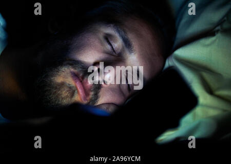 Bearded tired man in bed at night. He is trying to watch something on his phone but he is too tired to do so. Stock Photo