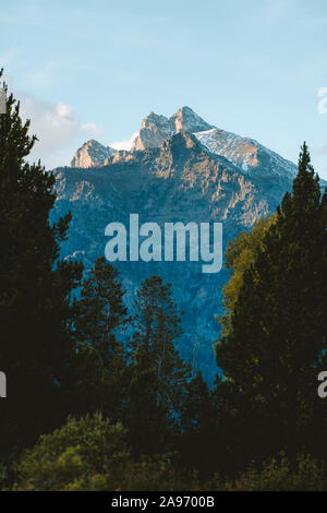 A mountain peak is illuminated by golden rays from the setting sun with trees in the foreground. Stock Photo