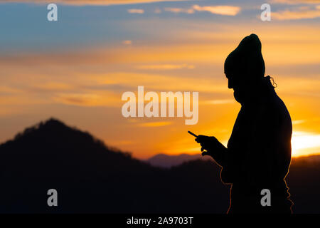 Silhouette of a male hiker with knit cap looking at his mobile phone on a beautiful sunset background. Hiking, modern technology and nature concepts. Stock Photo