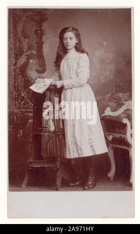 Early 1900's Edwardian studio portrait postcard of older attractive girl, teenage years, with long hair, wearing a pretty dress, boots and holding a magazine or book, from Allen Nield  Photographic studios in Leeds or Stockport studio, England, U.K. circa 1910. Stock Photo