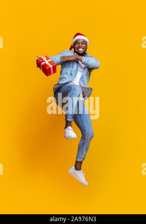 Christmas delivery concept. Positive african american guy jumping with Xmas present, hurry to deliver gift box in time. yellow background Stock Photo