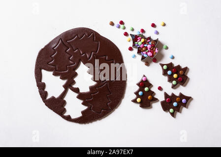Cocoa gingerbread dough and Christmas tree-shaped cookies decorated with colorful candies on white table. Making smarties gingerbread cookies. Xmas. Stock Photo
