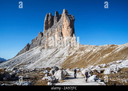 Two tourists walking under the south face of Tre Cime di Lavaredo / Drei Zinnen, mountain peaks in the Sesto / Sexten Dolomites, South Tyrol, Italy Stock Photo