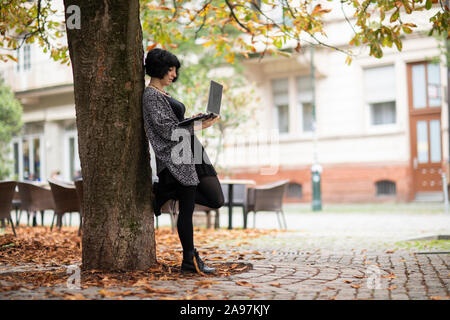 young woman with laptop outside working near a tree Stock Photo