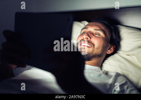 Handsome bearded smiling caucasian male lying in bed at night while watching something on his tablet. Stock Photo