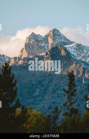 A mountain peak is illuminated by golden rays from the setting sun with trees in the foreground. Stock Photo