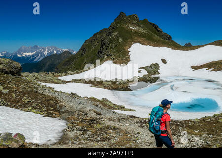 Young woman enjoying the view a frozen alpine lake. The ice shines in many different shades of blue. Lake is surrounded by tall mountains. No snow on Stock Photo