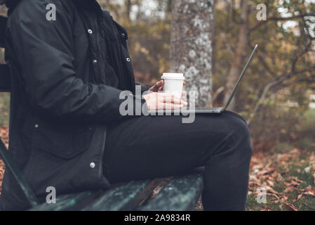 Young man drinking coffee with laptop in autumn park outdoors. Man with laptop and coffee in autumn park under fall foliage . Stock Photo