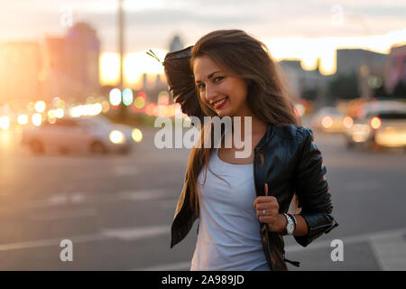 Beautiful young woman walking in the street with defocused city lights at the background. Hipster girl enjoying nightlife, fashion girl in leather Stock Photo