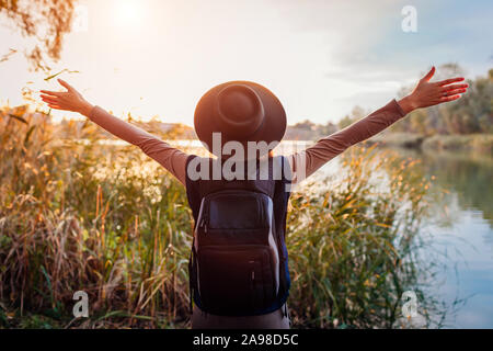 Traveler with backpack relaxing by autumn river at sunset. Young woman raised arms feeling free and happy Stock Photo