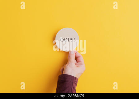 Male hand placing wooden cut circle with an Oops sign on it on bright yellow background. Stock Photo