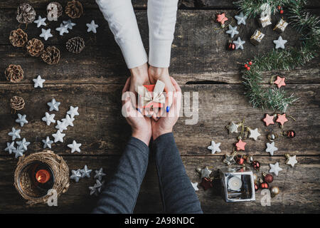 Top view of a male hands cupping female hands holding small red holiday gift box with golden bow. Over rustic wooden background full of Christmas orna Stock Photo