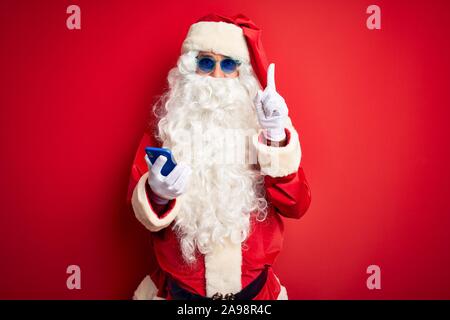 Senior man wearing Santa Claus costume using smartphone over isolated red background surprised with an idea or question pointing finger with happy fac Stock Photo