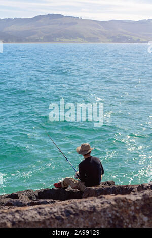 Unidentified man in white hat and dark shirt sitting on marina breakwater rocks fishing with a rod beside a sparkling blue ocean, at Apollo Bay Austra Stock Photo