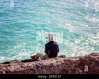 Unidentified man in white hat and dark shirt sitting on marina breakwater rocks fishing with a rod beside a sparkling blue ocean, at Apollo Bay Austra Stock Photo