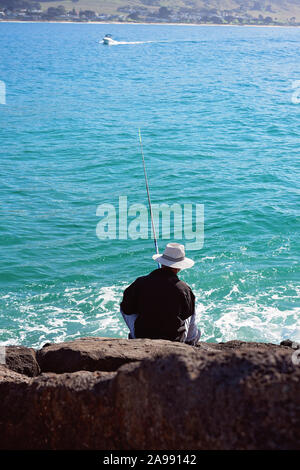 Unidentified man in white hat and dark shirt sitting on breakwater rocks fishing with a rod beside a sparkling blue ocean, at Apollo Bay Australia Stock Photo