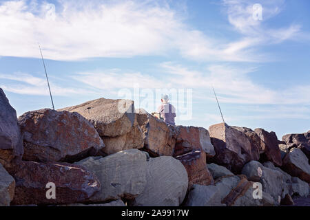 Unidentified man fishing with two rods out sitting on breakwater rocks with blue sky beyond, at Apollo Bay Australia Stock Photo