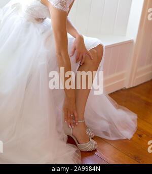 Bride fastens her show as she gets dressed for her wedding, framed by soft window light Stock Photo