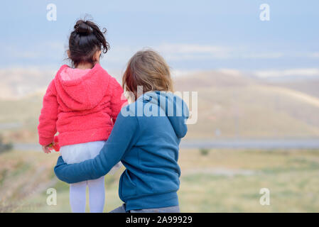 Young mother holding baby looking at amazing landscape.Family traveling in mountains. Back view mother and daughters standing and looking sunset lights. Stock Photo