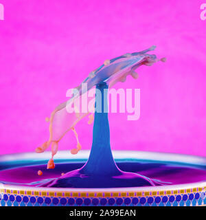 A colorful water drop with a blue spout and tumbling umbrella shape, on a hot pink background. Created with orange coloured milk dropped into blue liq Stock Photo