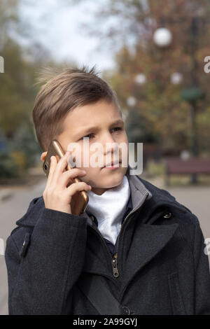 Portrait of a schoolboy dressed in a business suit talking on a cell phone, holding it in his hand near his ear. Front view with a serious expression Stock Photo