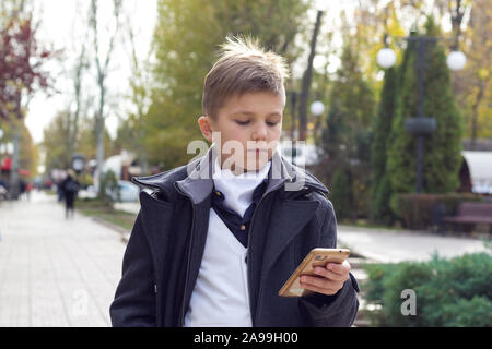 Schoolboy dressed in stylish business clothes holding smartphone in hand and watching on touch screen. Outdoors in park.Daytime in autumn. Child Stock Photo