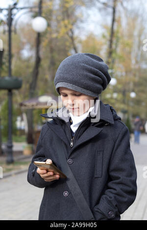 Schoolboy in hat holding smartphone in hand and dialing number on touch screen. Outdoors in park. Daytime in autumn. Child dressed in stylish business Stock Photo