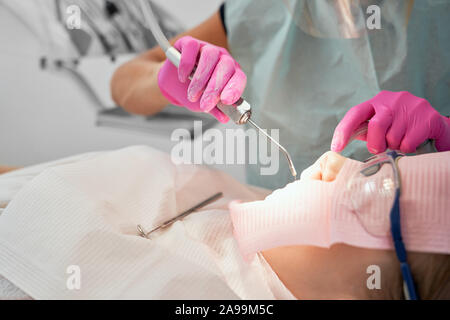 Patient of dental clinic sitting in dental chair with white bib and protective glasses. Dentist in rubber gloves holding tool, fitting teeth. Doctor doing procedure with dental curing UV light. Stock Photo