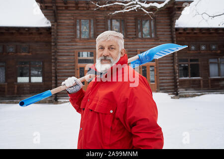 Portrait of cleaner with snow shovel on background of wooden house in winter. Elderly man in warm red jacket holding shovel on shoulder, looking at camera. Season of removing snow. Stock Photo