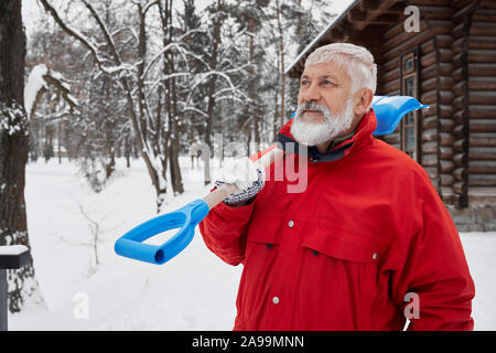 Elderly man standing near wooden house and holding snow shovel on shoulder. Bearded cleaner wearing in red jacket looking away. Concept manual removing snow after snowstorm. Stock Photo