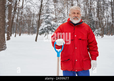 Janitor wearing warm jacket when cleaning snow. Elderly man of city service holding snow shovel and posing outdoors in park. Concept of snow removal after snowstorm. Stock Photo