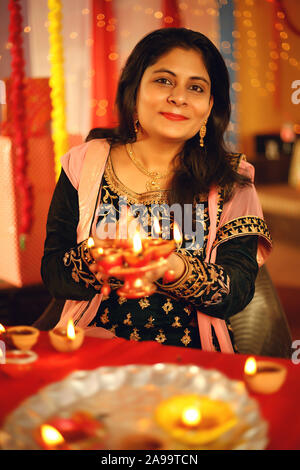 A beautiful young Indian woman in traditional sari dress holding an oil lamp light or Diya within decorative background on the occasion of Diwali Stock Photo