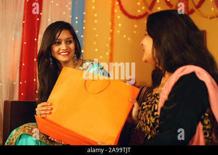 Cute young Indian woman wearing traditional ethnic clothes giving gift to a woman, over festive bokeh background. Stock Photo