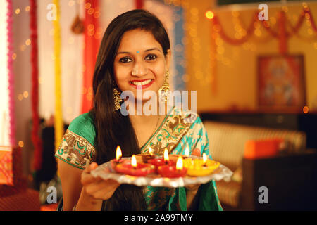 A beautiful young Indian woman in traditional sari dress holding a oil lamp light or diya with in decorative background on the occasion of Diwali. Stock Photo