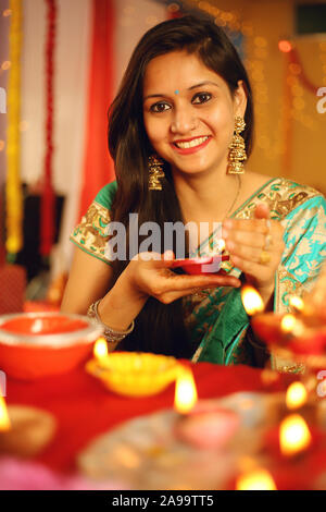 A beautiful young Indian woman in traditional sari dress holding an oil lamp light or Diya within the decorative background on the occasion of Diwali. Stock Photo