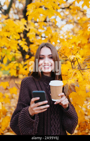 happy positive cute young student girl in autumn park using mobile phone. and drinking coffee Stock Photo