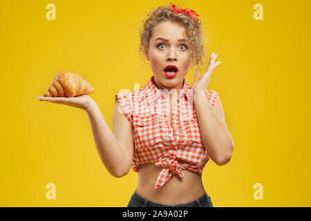 Attractive lady with blond wavy hair looks shocked because she decided to eat delicious croissant during her diet period. Concept of unhealthy food and will power Stock Photo