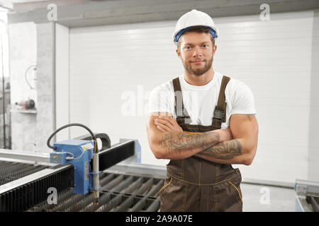 Front view of handsome man in uniform and helmet standing with folded arms, looking at camera and smiling. Male mechanic working with cnc on metal factory. Concept of machinery. Stock Photo